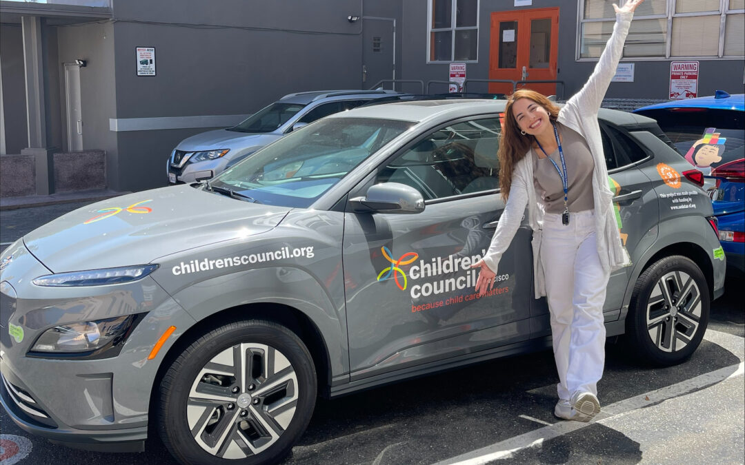 A smiling Children's Council staff member stands in front of Children's Council's new electric vehicle with their arms open.